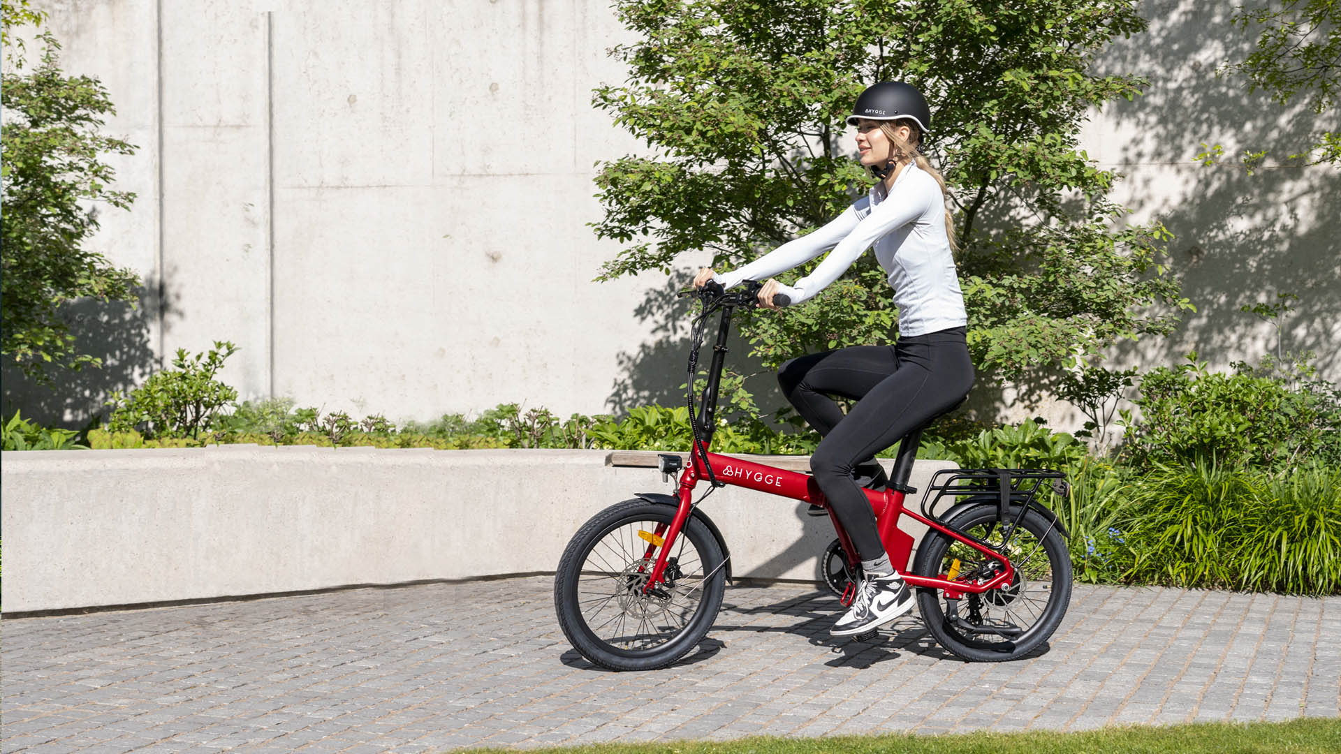 Woman riding sangria red Hygge Virum lightweight folding e-bike in urban setting with greenery